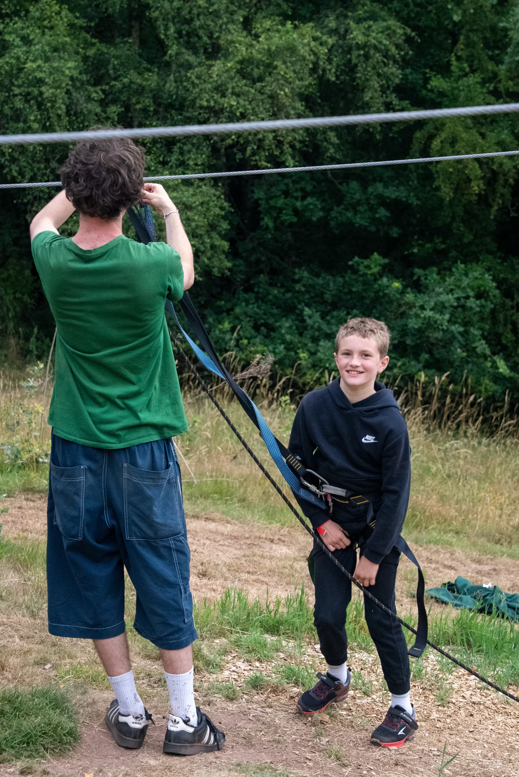 Friendly outdoor adventure park guide in uniform helping a smiling young girl adjust her safety harness before a zip line ride, supportive encouraging instructor, sunny outdoor nature park, candid professional photography